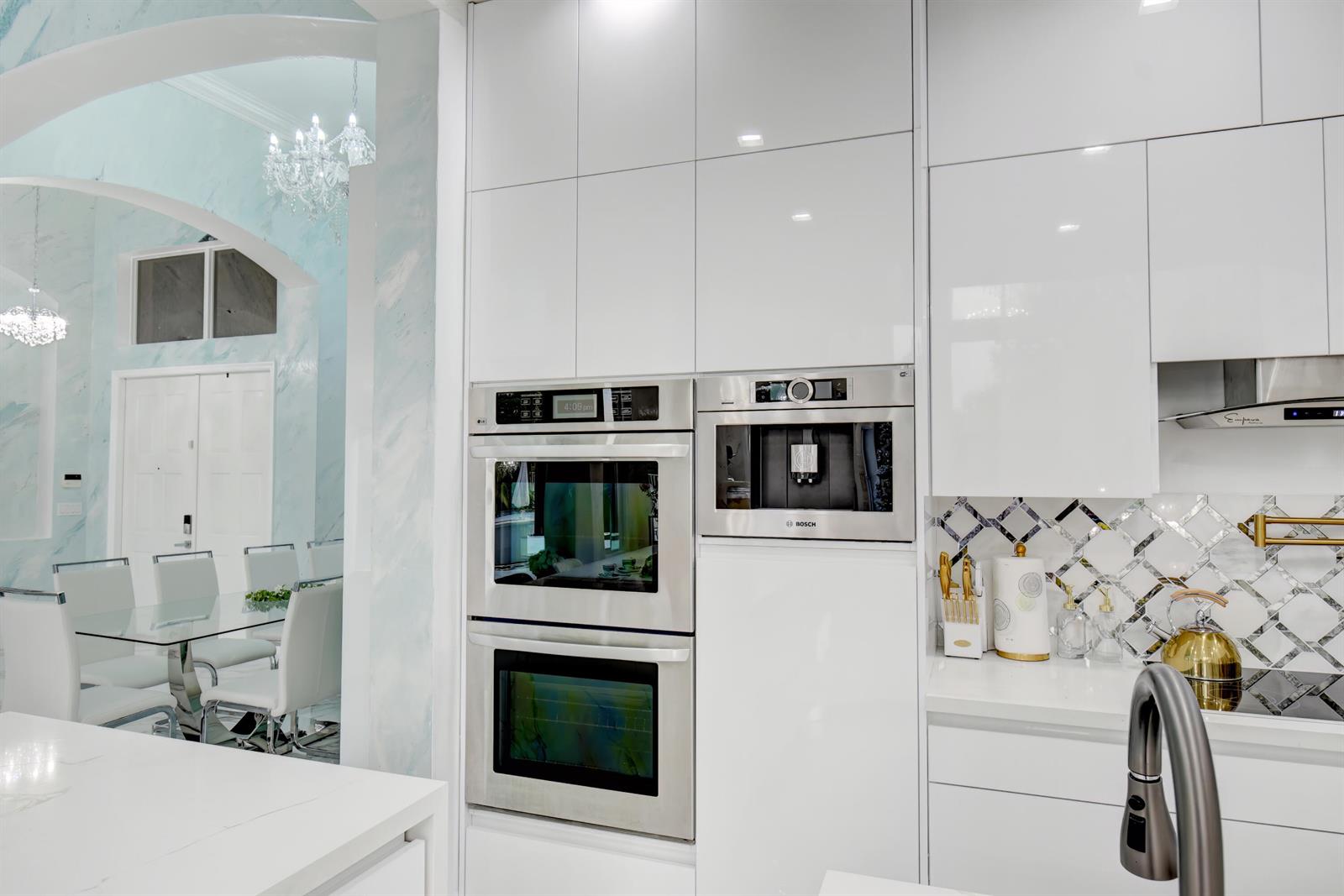 A modern kitchen features white glossy cabinetry, stainless steel built-in ovens, and a coffee machine — 11910 Osprey Point Circle, Wellington FL 33449, WELLINGTON, FL