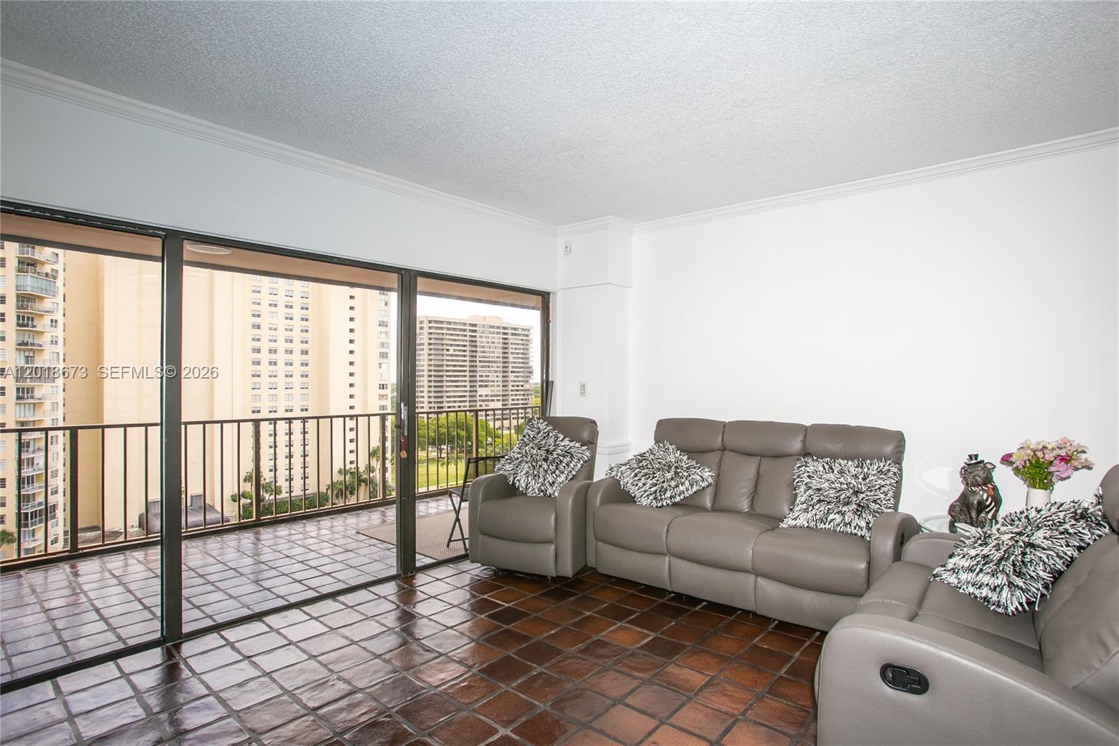 A living room features dark tile flooring, gray leather furniture, and a balcony with city views — 1800 NE 114th St # 1207, Miami FL 33181, MIAMI, FL