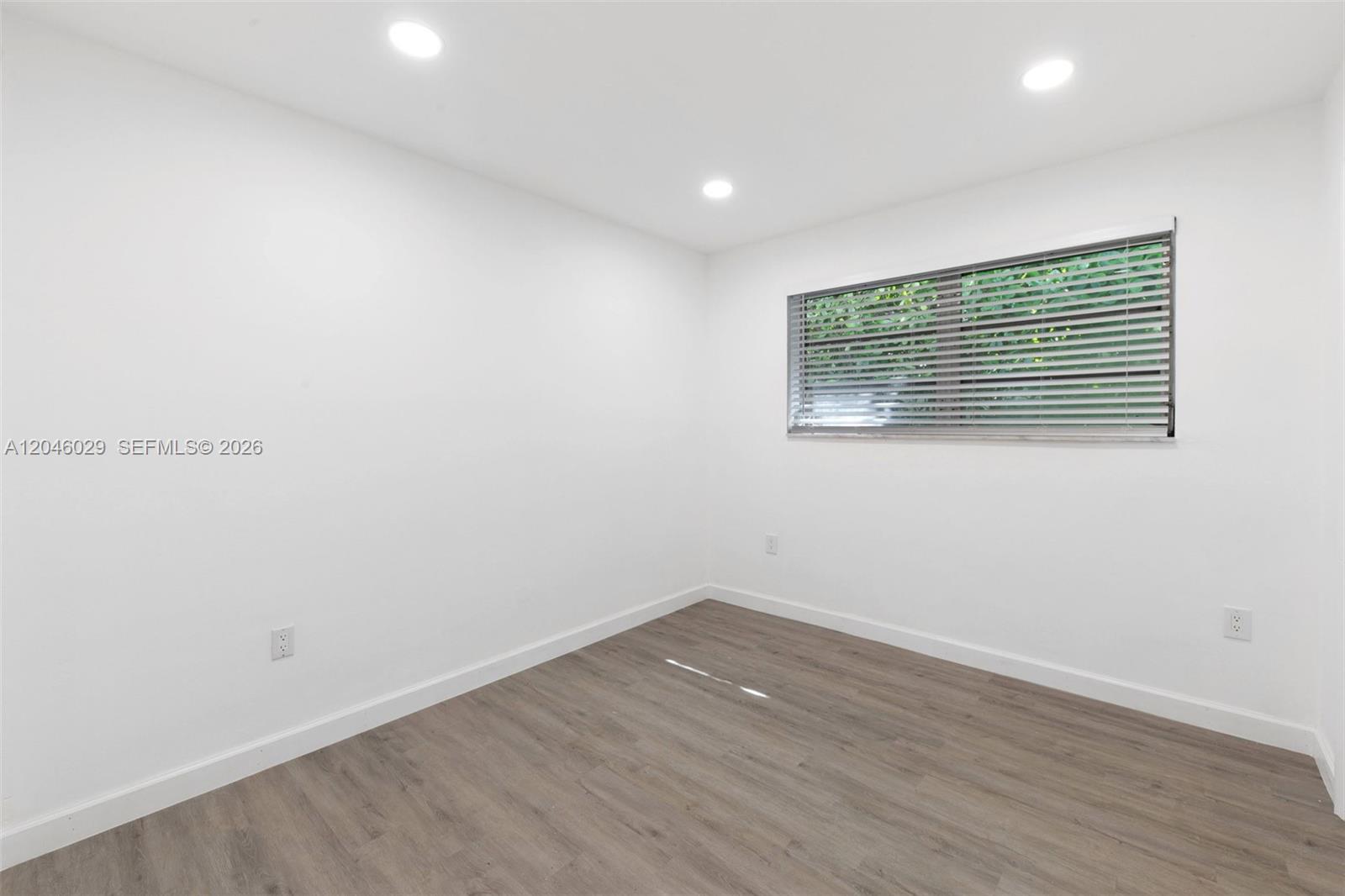 An empty room features white walls, recessed lighting, a window with blinds, and wood-look flooring — 945 NE 176th St, Miami FL 33162, MIAMI, FL