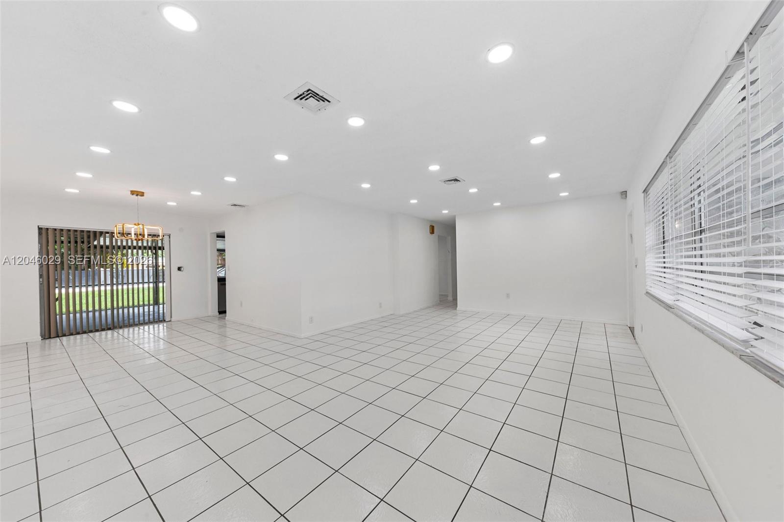 A bright, empty room features white tiled floors, recessed lighting, and large windows with blinds — 945 NE 176th St, Miami FL 33162, MIAMI, FL