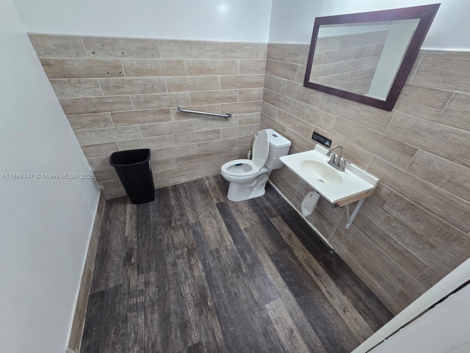 A small bathroom features wood-look tile walls, dark flooring, a toilet, and a wall-mounted sink — 2501 N State Road 7 # 2503, Margate FL 33063, MARGATE, FL