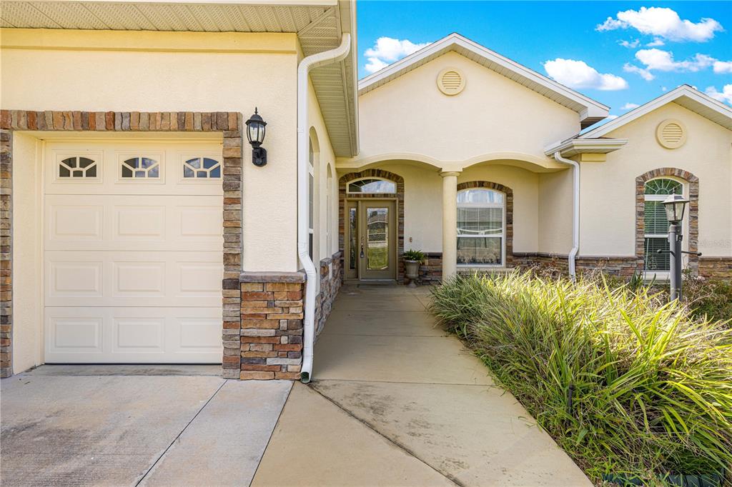 A beige house exterior features a stone-trimmed garage, a covered entryway, and a concrete walkway — 17306 SE 112TH CT ROAD, SUMMERFIELD, FL