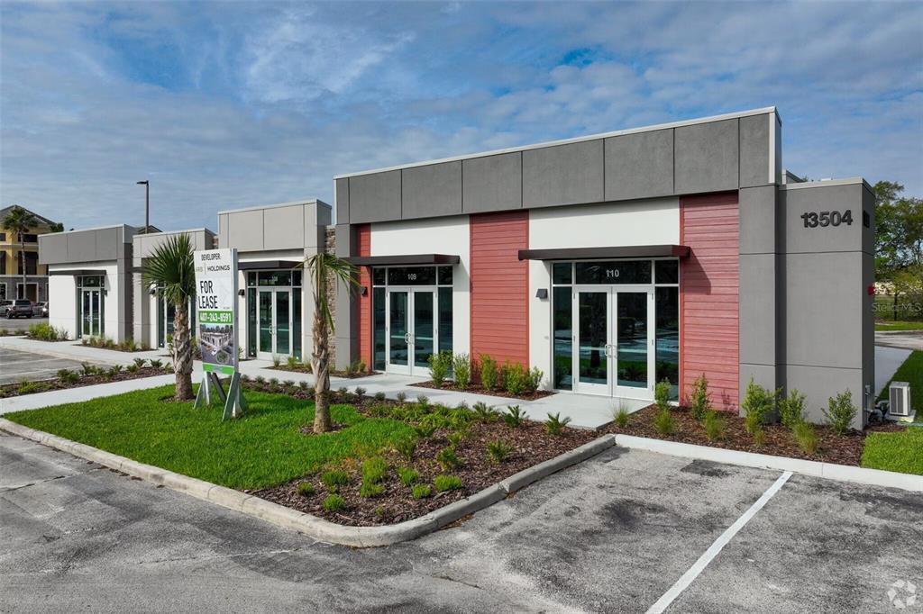 A modern commercial building exterior features gray panels, red siding, and glass double-door entrances — 13504 VILLAGE PARK DRIVE UNIT 103, ORLANDO, FL