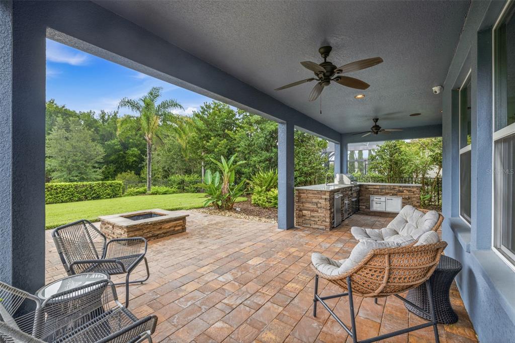 A covered patio features a stone fire pit, outdoor kitchen, and seating area overlooking a yard — 31321 DRISCOLL DRIVE, WESLEY CHAPEL, FL