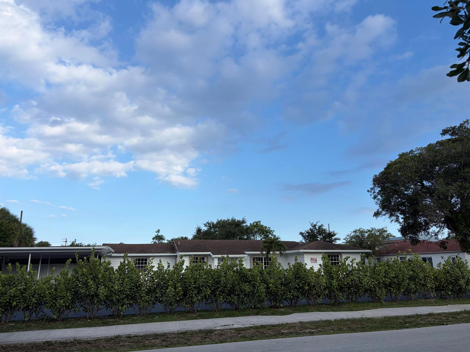 A white single-story house with a brown roof sits behind a long, dense green hedge — 202 Palm Court, Delray Beach FL 33444, DELRAY BEACH, FL