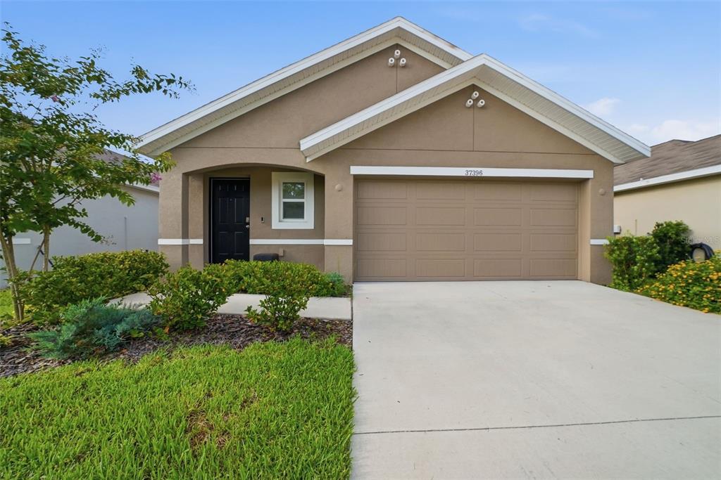 A single-story suburban house with a tan stucco exterior, a two-car garage, and a driveway — 37396 SHARKS EYE LANE, ZEPHYRHILLS, FL