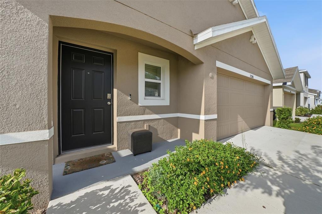A tan stucco house exterior features a black front door, a garage, and a garden bed — 37396 SHARKS EYE LANE, ZEPHYRHILLS, FL