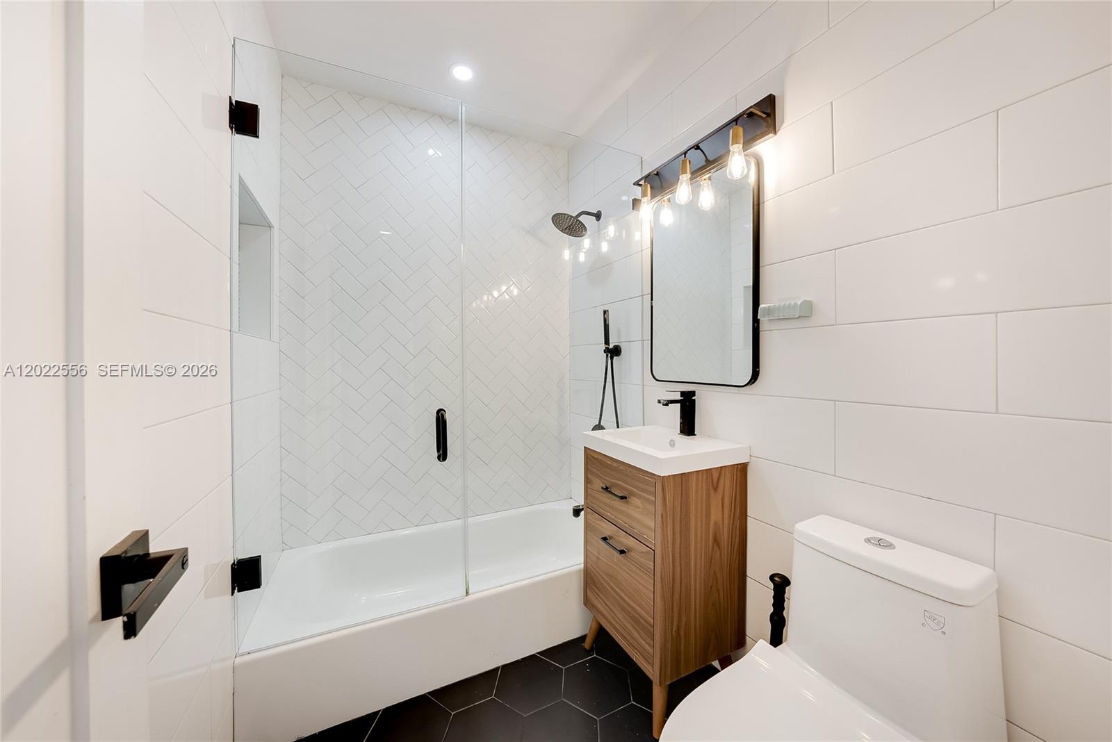 A modern bathroom features a white subway-tiled shower, a wood vanity, and black hexagonal flooring — 716 SW 5th Ave, Hallandale Beach FL 33009, HALLANDALE BEACH, FL