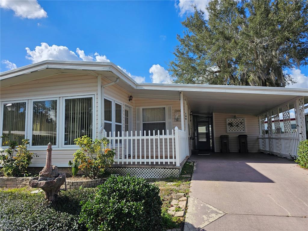 A light-colored mobile home features a white picket fence, a carport, and a small fountain — 104 IXORA WAY, LEESBURG, FL