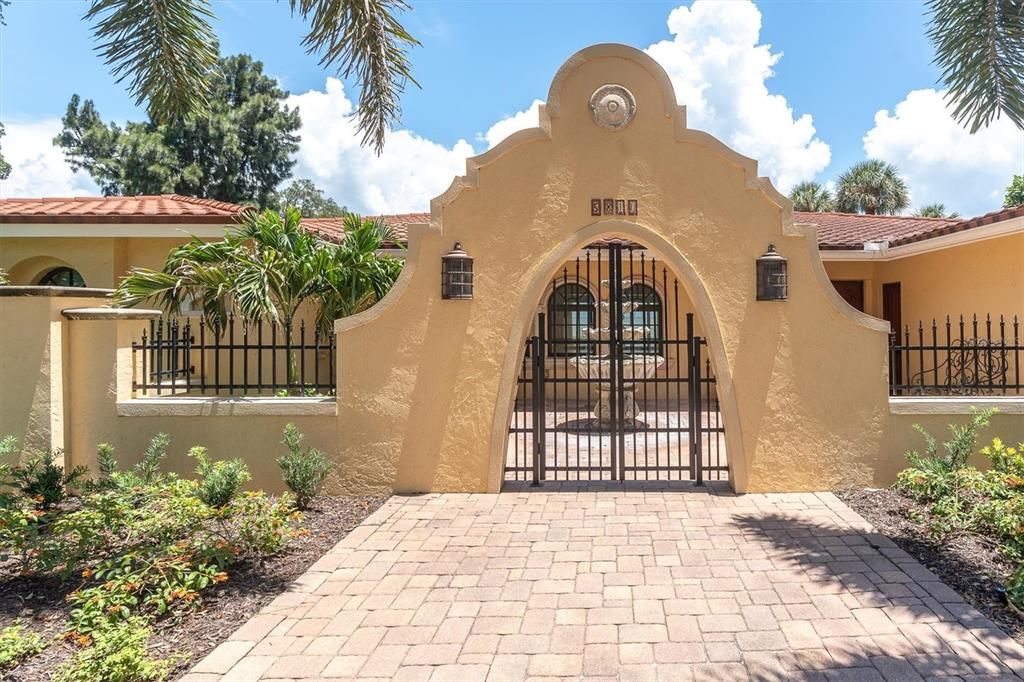 A stucco wall with a decorative arched gate and iron fencing surrounds a paved courtyard — 5810 BAHAMA SHORES DRIVE S, ST PETERSBURG, FL