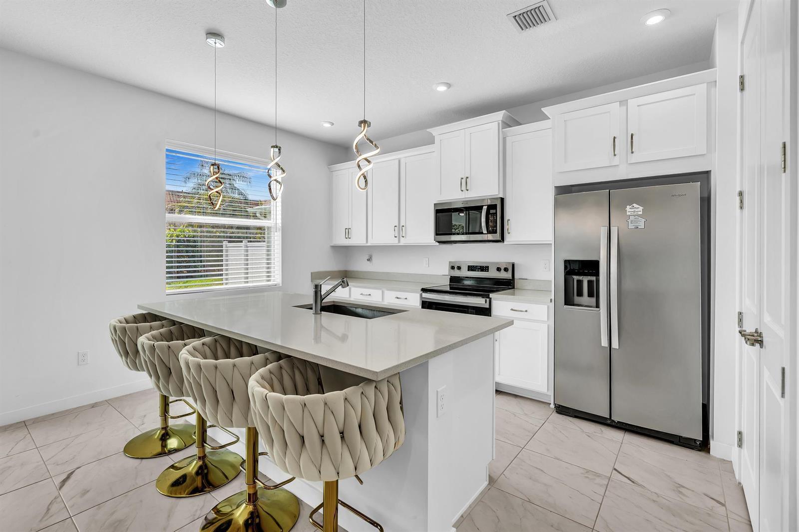 A modern kitchen features white cabinets, stainless steel appliances, a quartz island, and gold-based stools — 7268 Lenora Lane, Lake Worth FL 33467, LAKE WORTH, FL