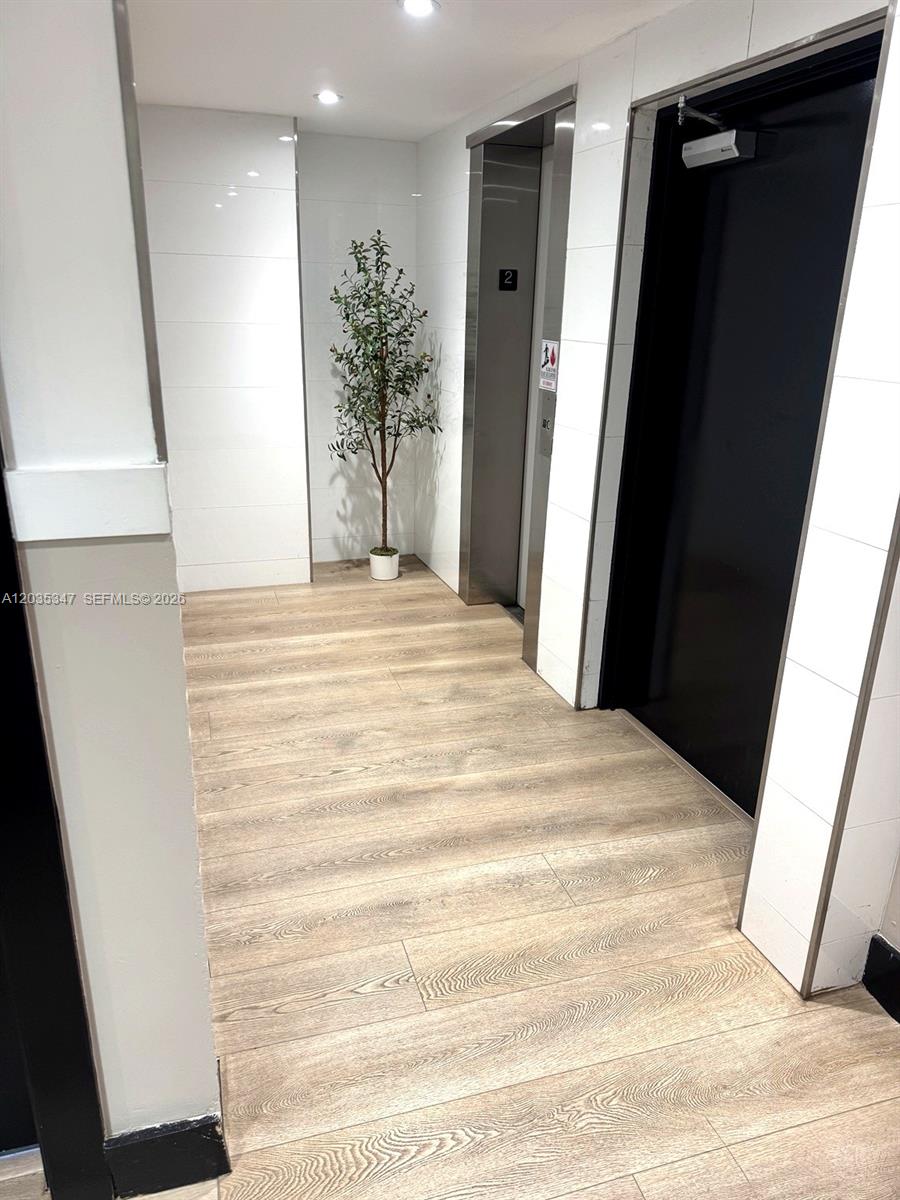 A hallway features wood-look flooring, white tiled walls, an elevator, and a potted plant — 9491 Harding Ave # 205, Surfside FL 33154, SURFSIDE, FL