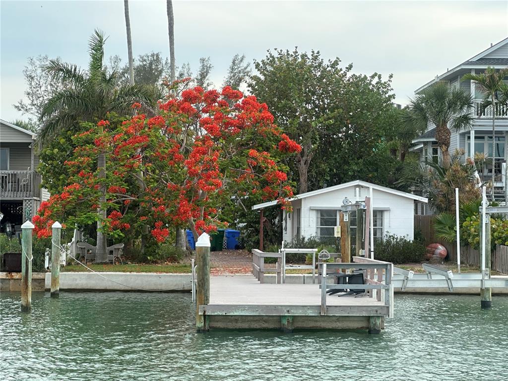 A wooden dock extends into a canal in front of a house with a flowering tree — 8405 BAYSHORE DRIVE, TREASURE ISLAND, FL