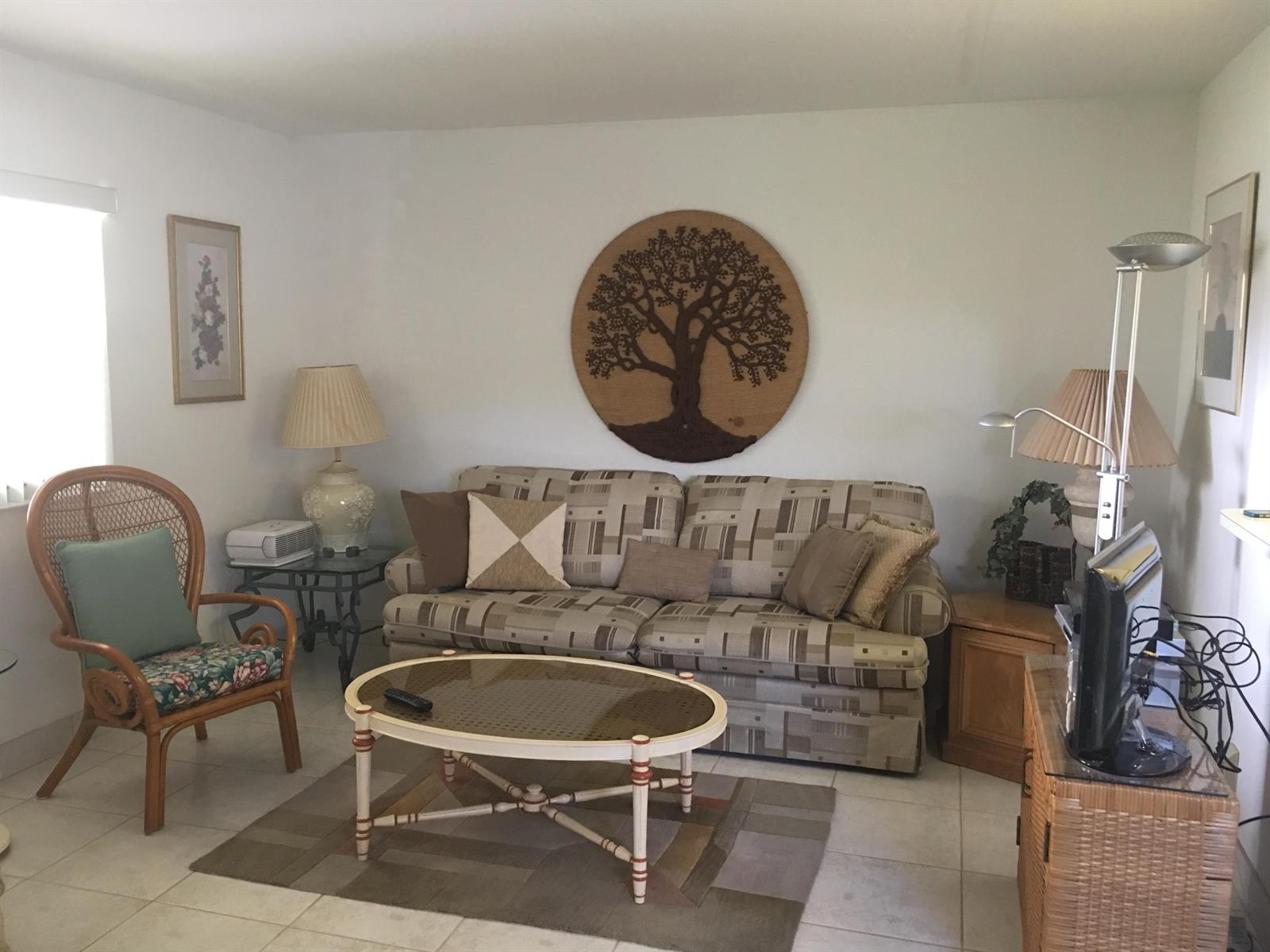 A living room features a patterned sofa, a round tree wall art, and a wicker chair — 99 Saxony C, Delray Beach FL 33446, DELRAY BEACH, FL
