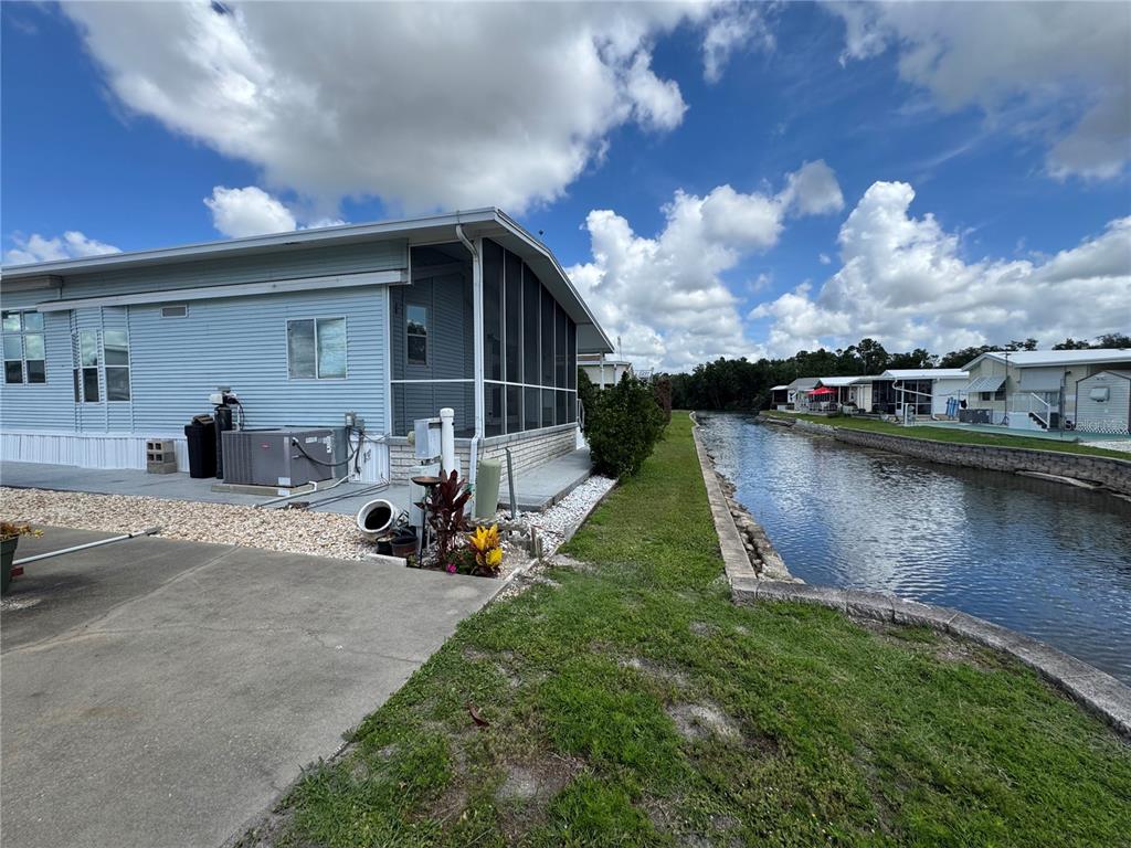 A light blue mobile home sits beside a narrow canal under a cloudy blue sky — 35151 DANNY DRIVE Lot 114, ZEPHYRHILLS, FL