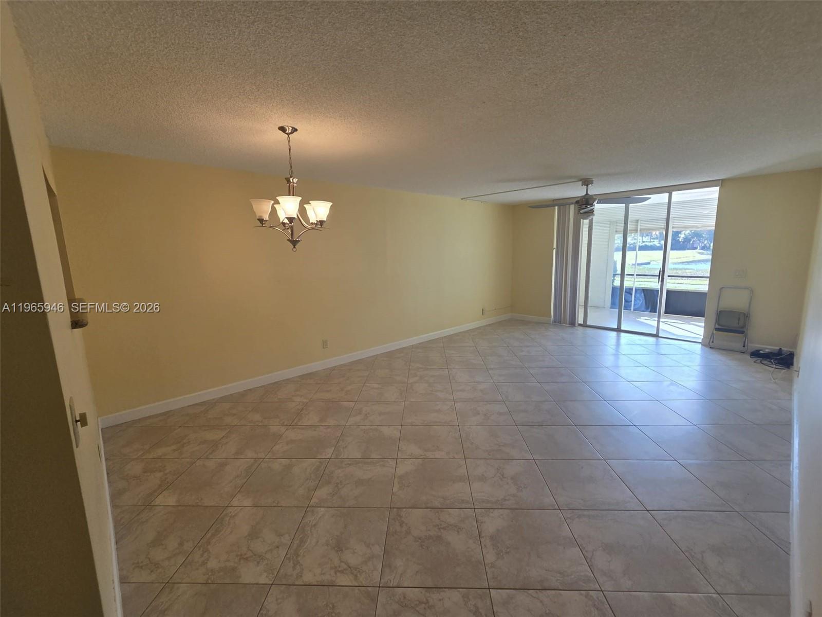 An empty living room features light-colored tile flooring, yellow walls, a chandelier, and sliding glass doors — 6675 S Oriole Blvd # 1040, Delray Beach FL 33446, DELRAY BEACH, FL