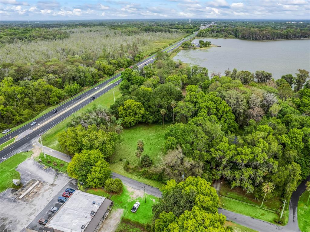 An aerial view shows a grassy lot bordered by a highway, trees, and a lake — S US HWY 27 & HOWARD RD, LEESBURG, FL