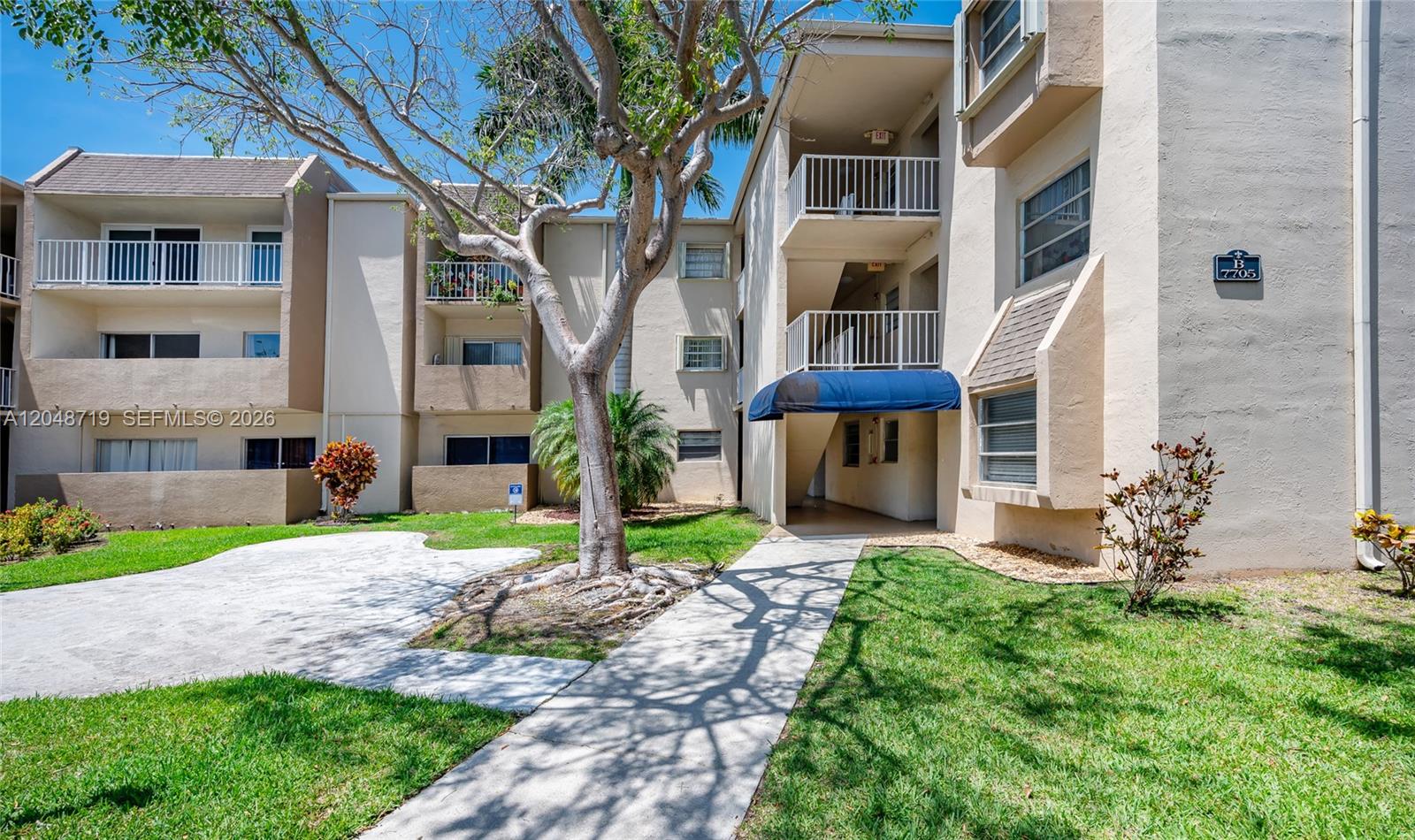 A multi-story apartment building exterior features tan stucco walls, balconies, and a concrete walkway — 7705 SW 86th St # 114, Miami FL 33143, MIAMI, FL