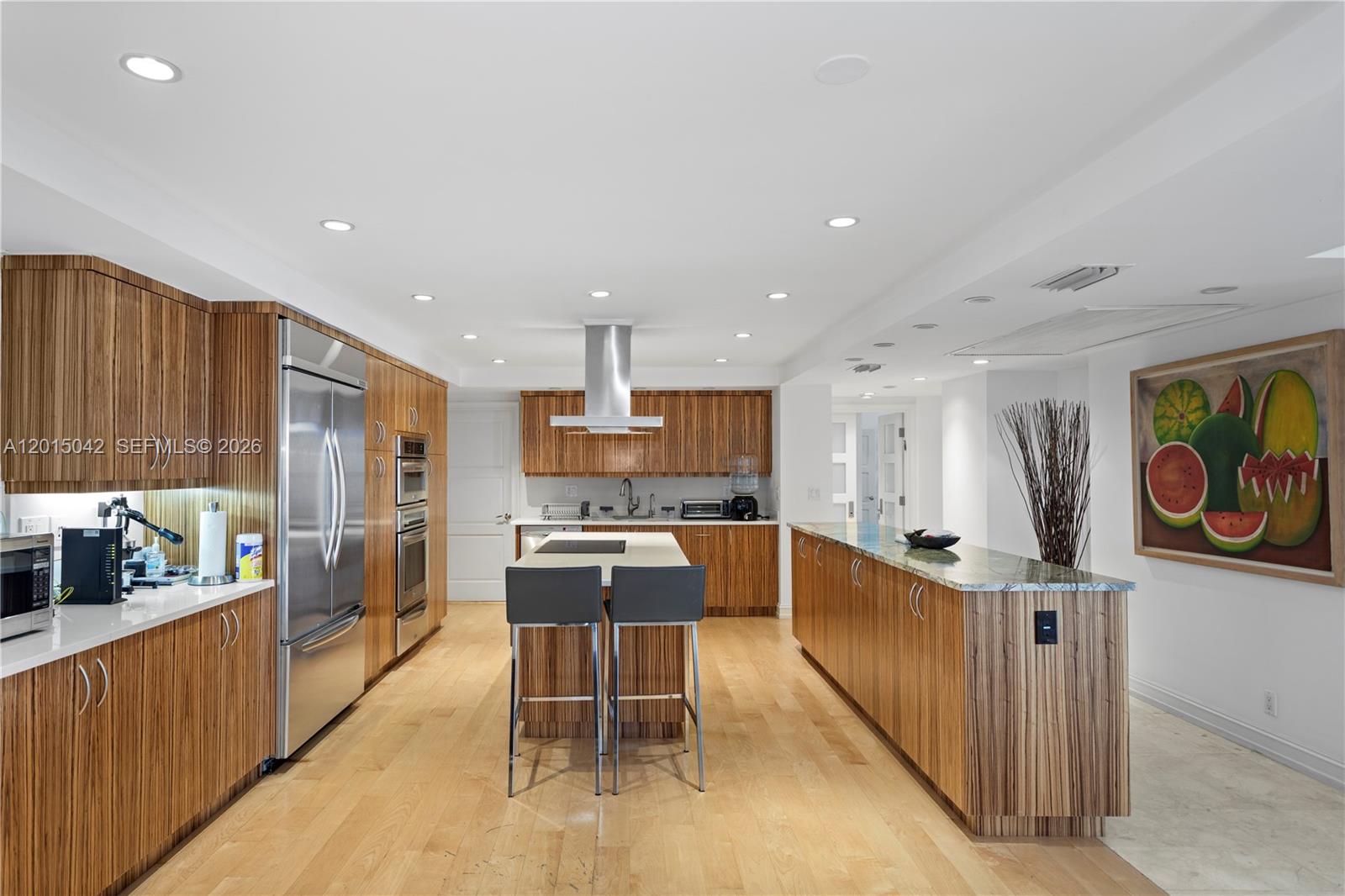 A modern kitchen features wood-grain cabinetry, a central island with bar stools, and stainless appliances — 10155 Collins Ave # 1105, Bal Harbour FL 33154, BAL HARBOUR, FL
