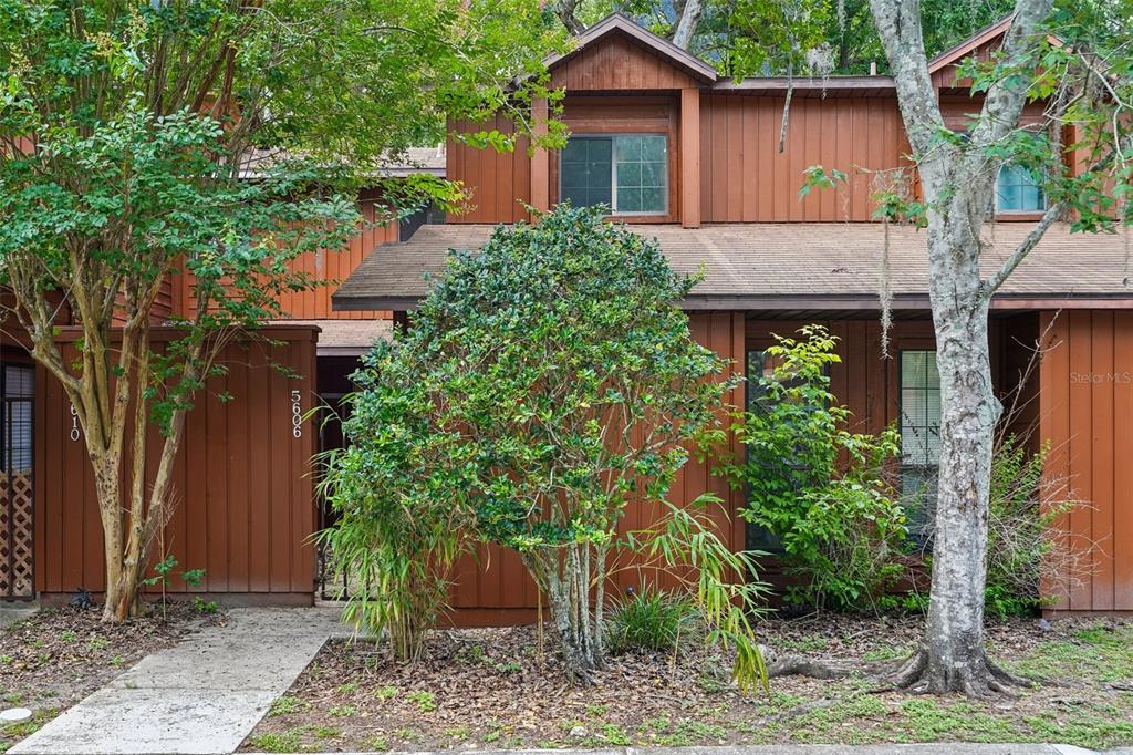 A two-story brown wood-sided townhouse features a front walkway, a large bush, and trees — 5606 SW 8TH PLACE, GAINESVILLE, FL