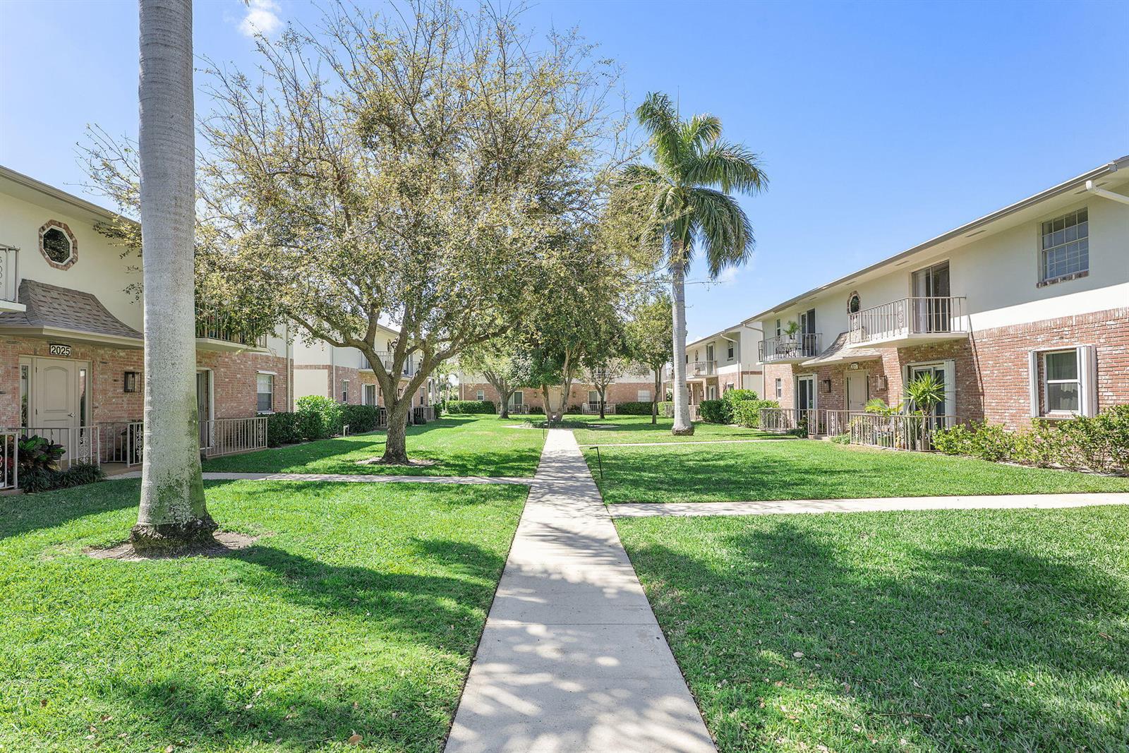 A paved walkway leads through a grassy courtyard between two-story apartment buildings with palm trees — 2015 S Seacrest Boulevard # C, Boynton Beach FL 33435, BOYNTON BEACH, FL