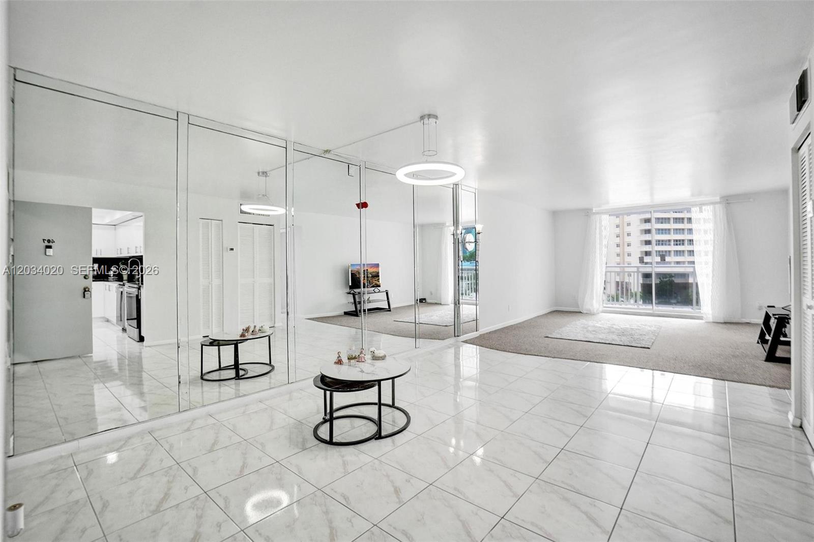 A living room with white tile floors, mirrored walls, and a carpeted area near windows — 1849 S Ocean Dr # 407, Hallandale Beach FL 33009, HALLANDALE BEACH, FL