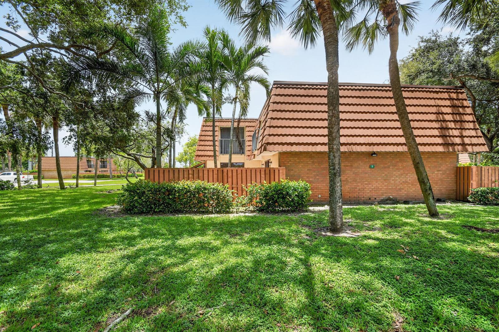 A brick townhouse with a brown tiled roof sits behind a wooden fence and lawn — 135 Heritage Way, West Palm Beach FL 33407, WEST PALM BEACH, FL