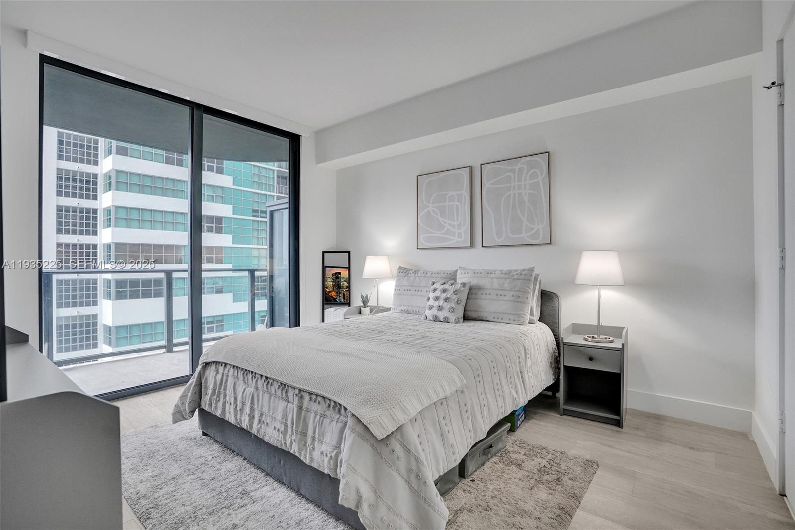 A modern bedroom features a bed with grey bedding, two bedside lamps, and floor-to-ceiling windows — 121 NE 34th St # 902, Miami FL 33137, MIAMI, FL