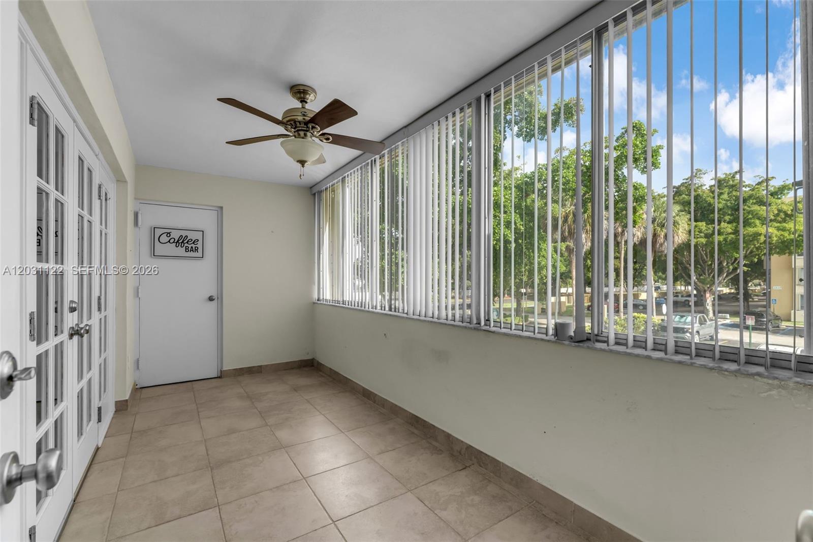 This enclosed balcony features tile flooring, a ceiling fan, and vertical blinds overlooking trees — 6911 SW 147th Ave # 2H, Miami FL 33193, MIAMI, FL