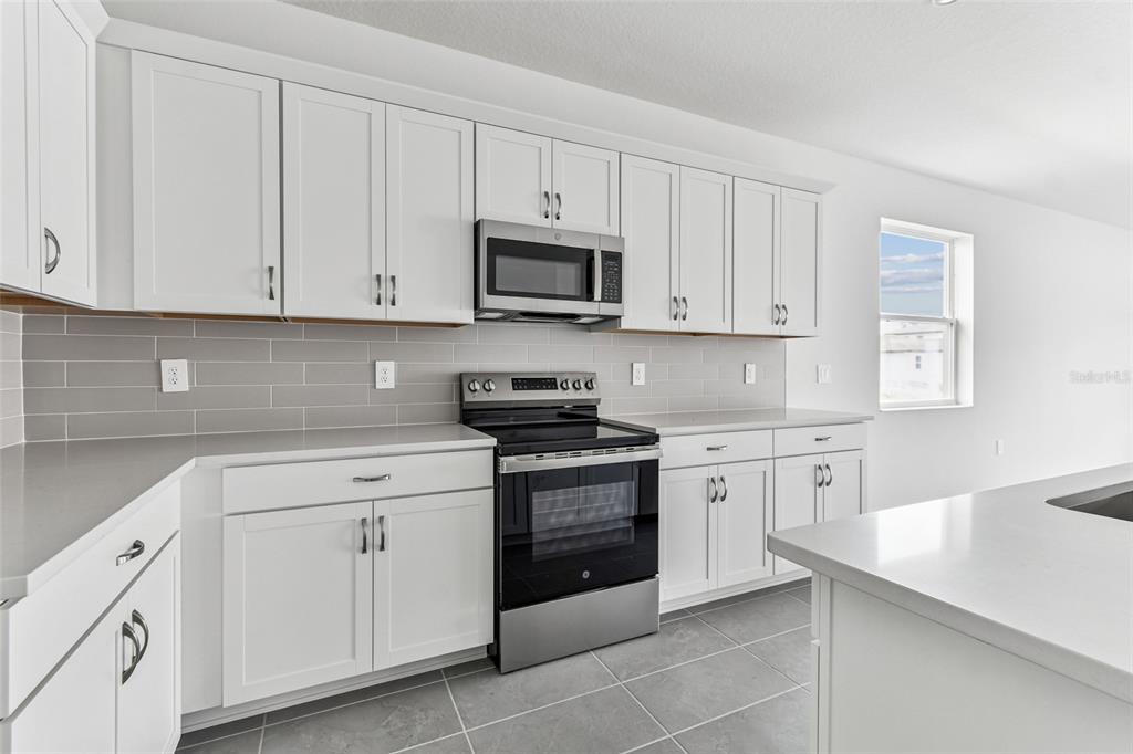 A modern kitchen features white cabinets, stainless steel appliances, gray tile backsplash, and quartz countertops — 9122 OPTIMIST WAY, PALMETTO, FL