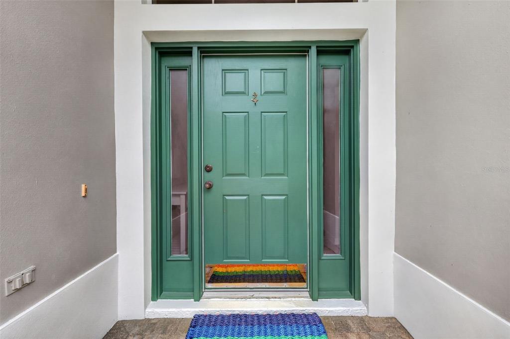 A green front door with side windows and a colorful mat sits between gray walls — 1218 CAMERO DRIVE, LADY LAKE, FL