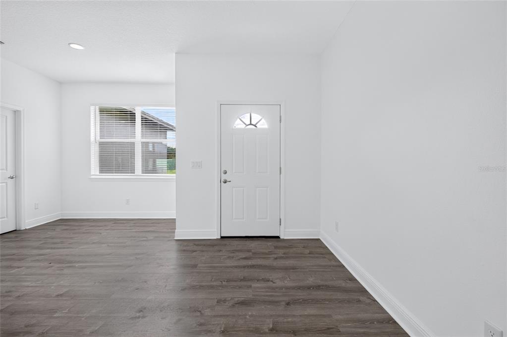 A bright, empty room features white walls, dark wood-look flooring, and a front entry door — 1400 SEBRING WAY, POINCIANA, FL