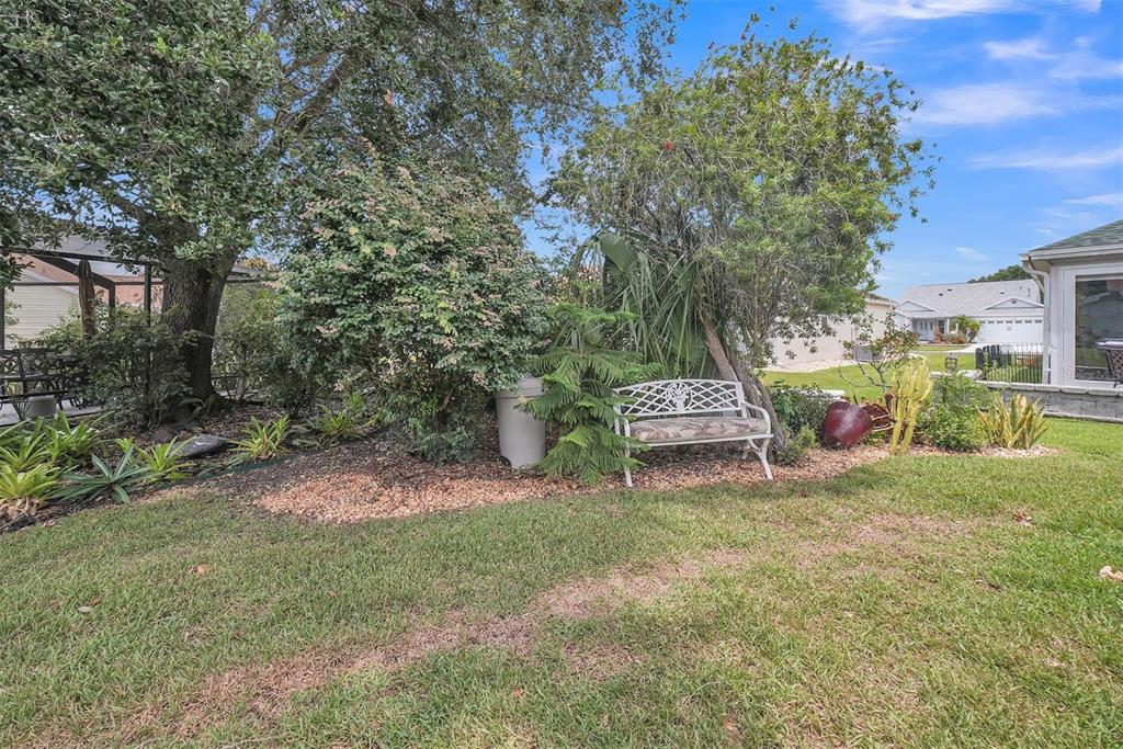 A white metal bench sits in a landscaped garden bed next to a large tree — 8371 SE 168TH TRINITY PLACE, THE VILLAGES, FL
