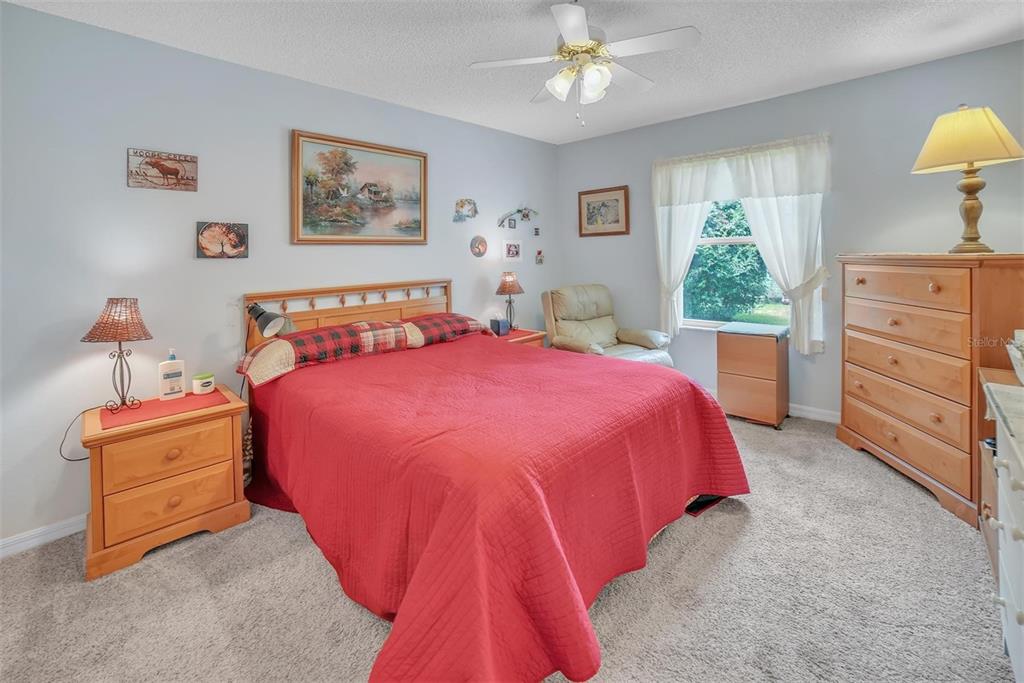 A bedroom features a red bedspread, light wood furniture, carpeted flooring, and a ceiling fan — 8371 SE 168TH TRINITY PLACE, THE VILLAGES, FL