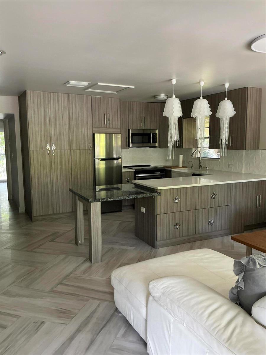 A modern kitchen features wood-grain cabinetry, a granite-topped island, and three white jellyfish-style pendant lights — 40 Ventnor Drive, Deerfield Beach FL 33442, DEERFIELD BEACH, FL