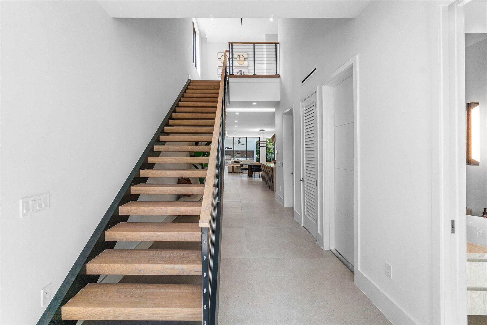 A modern hallway features a floating wooden staircase with a black metal railing and tile flooring — 810 Palm Trail, Delray Beach FL 33483, DELRAY BEACH, FL