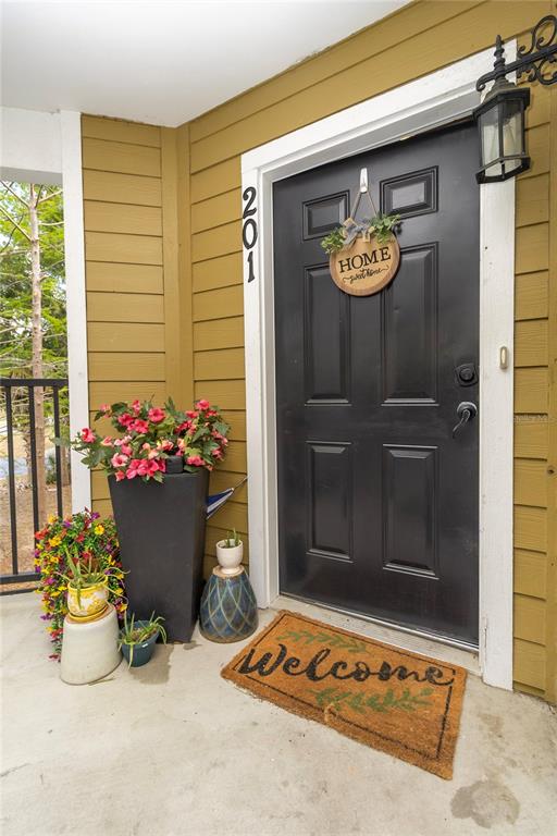 A black front door with a welcome mat and potted plants sits on a porch — 4215 SW 31ST DRIVE UNIT 201, GAINESVILLE, FL