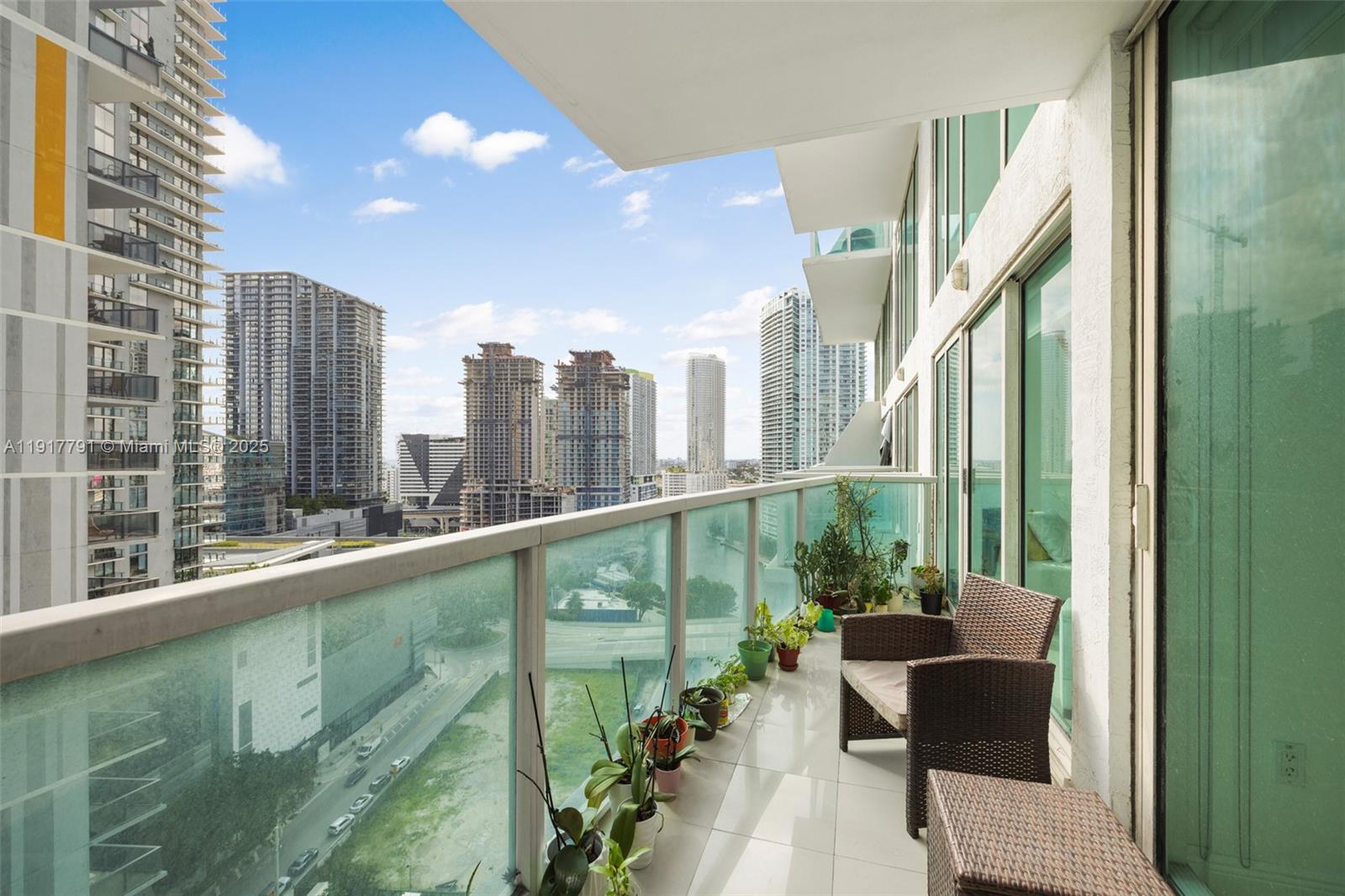 A high-rise balcony features a glass railing, potted plants, and outdoor furniture overlooking city buildings — 41 SE 5th St # 1210, Miami FL 33131, MIAMI, FL