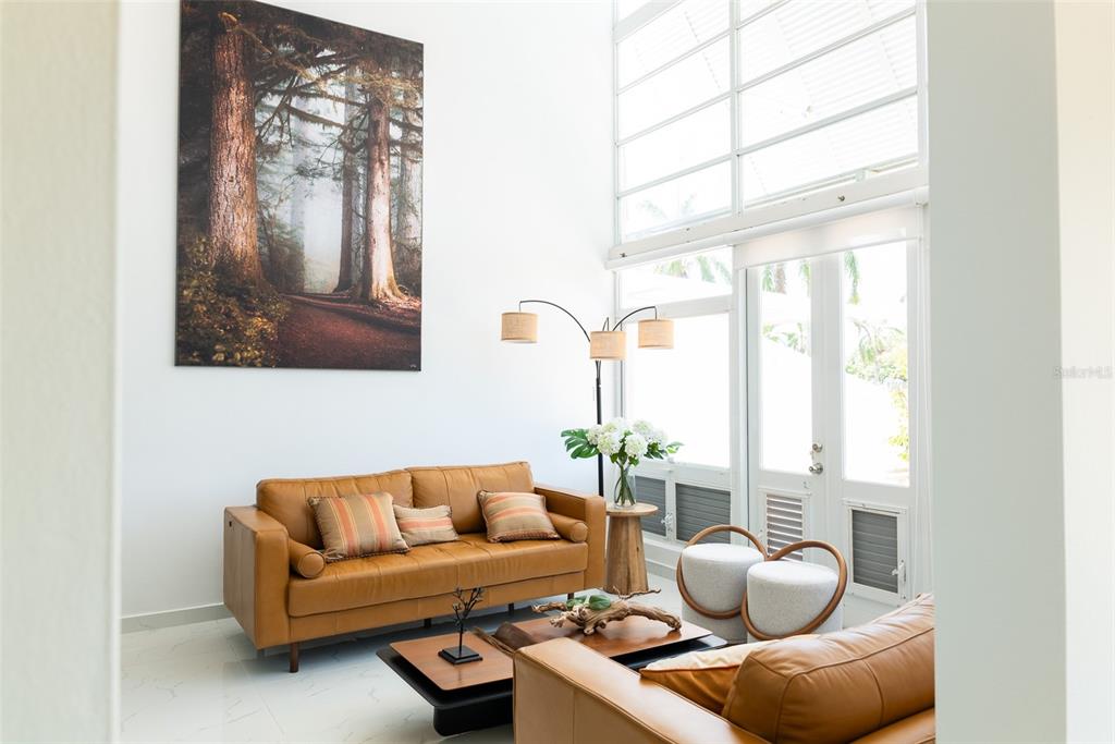 A living room features a tan leather sofa, tall windows, and a large forest wall art — 91 VILLAS DE GOLF EAST, DORADO, PR