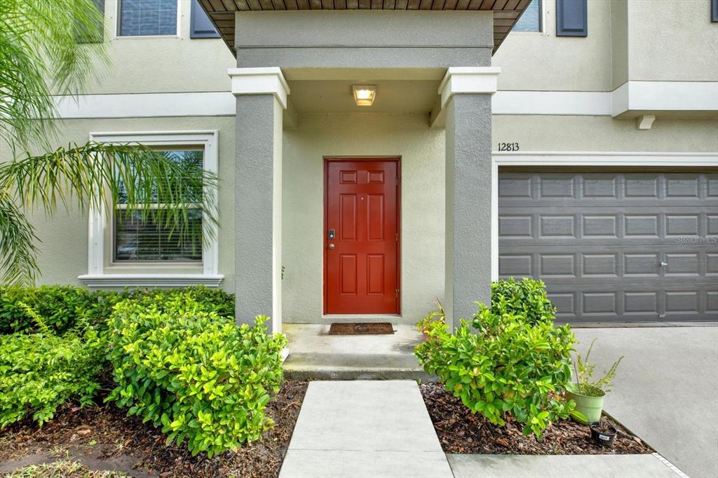 A red front door is centered under a covered entryway next to a gray garage — 12813 WILDFLOWER MEADOW DRIVE, RIVERVIEW, FL