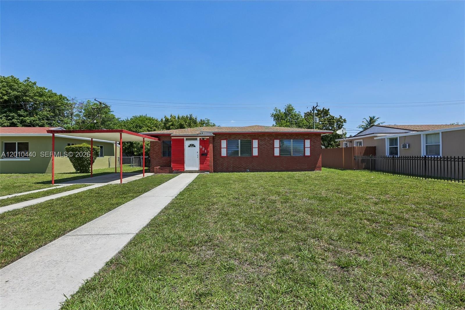 A single-story brick house features a red front entrance, white shutters, and a metal carport — 921 NE 159th St, North Miami Beach FL 33162, NORTH MIAMI BEACH, FL
