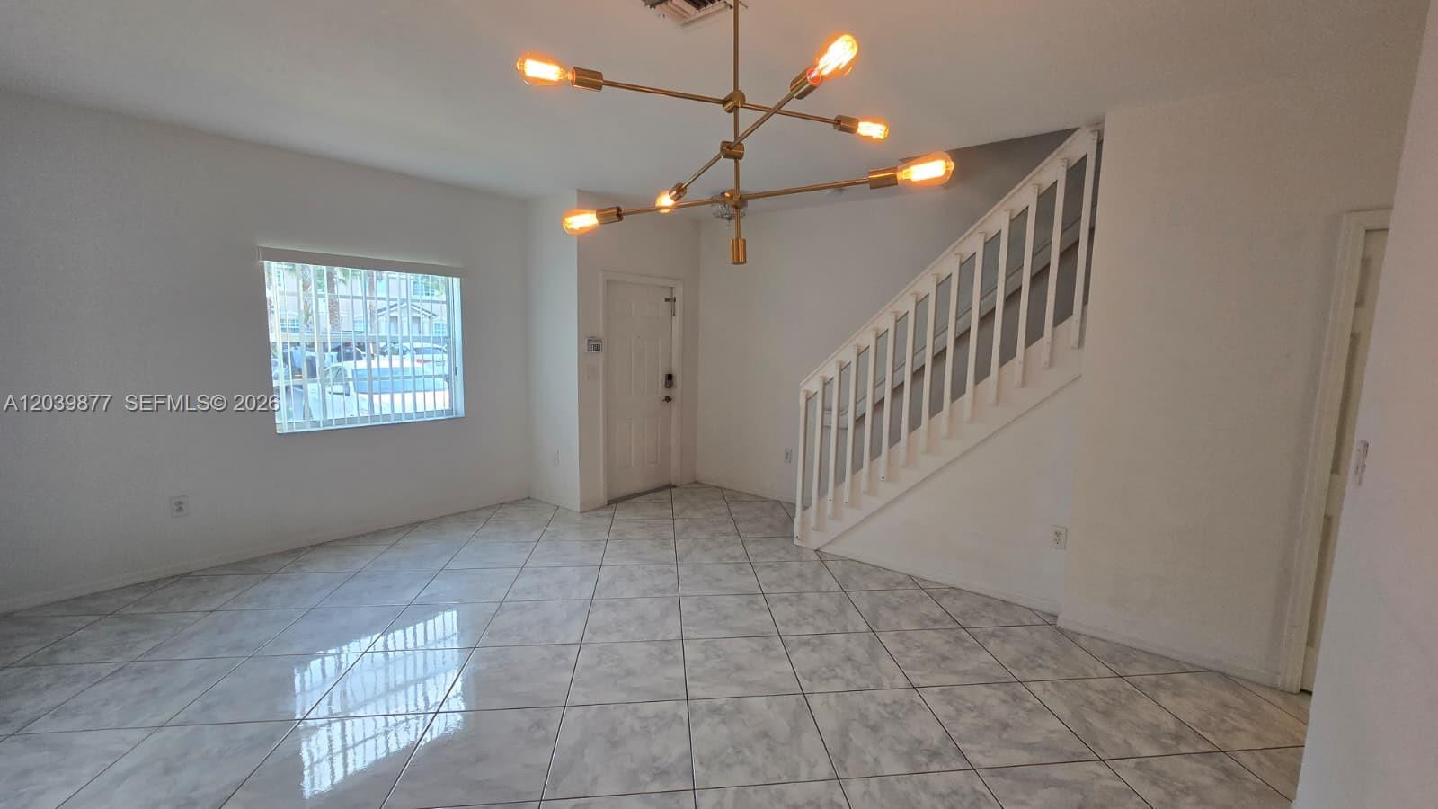 An empty room features white walls, tile flooring, a modern chandelier, and a staircase — 5791 NW 116th Ave # 101, Doral FL 33178, DORAL, FL