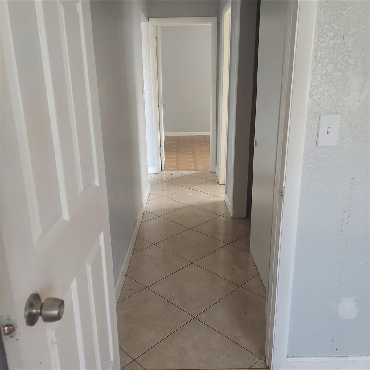 A hallway with light-colored tile flooring leads to a doorway showing a room with wood flooring — LAUDERDALE LAKES, FL