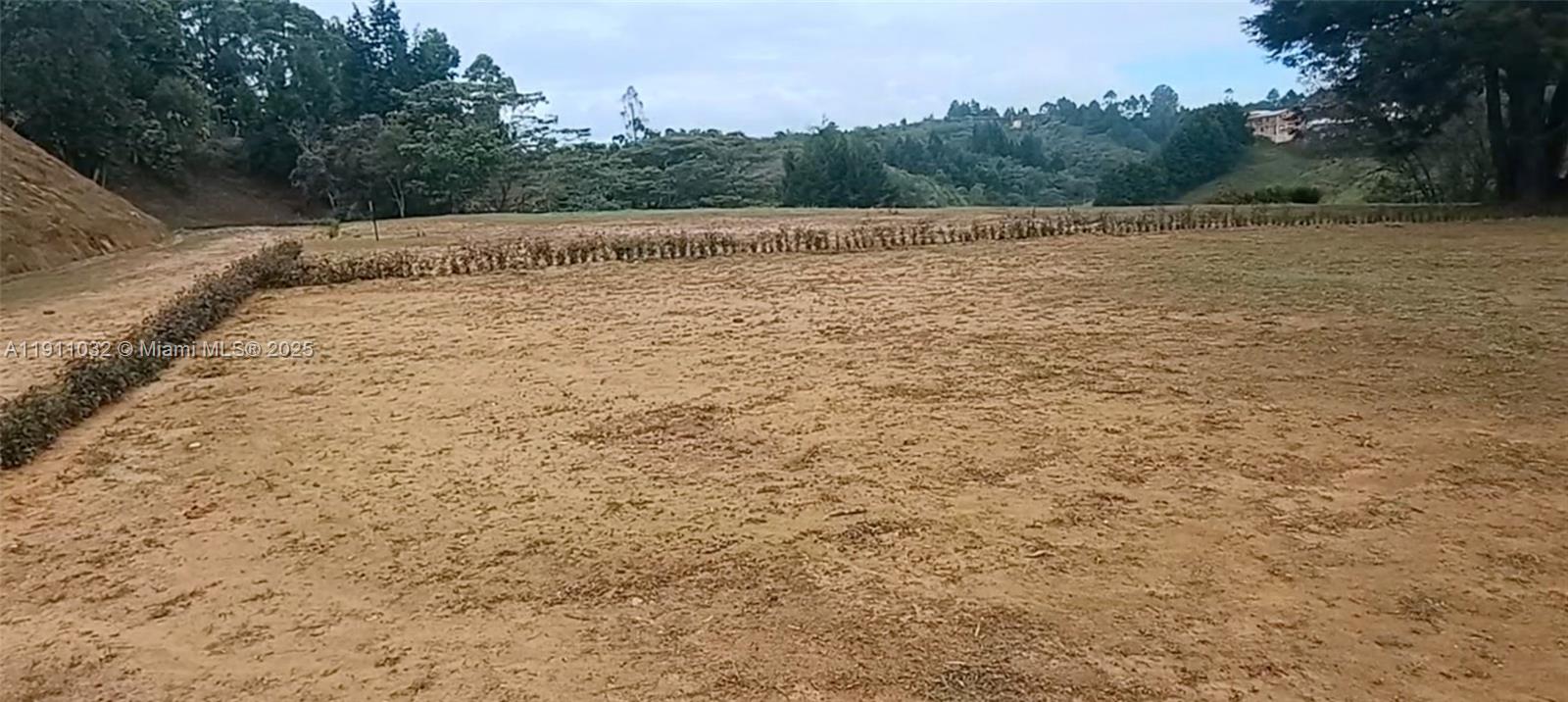 A large, cleared, dirt-covered plot of land sits in front of a hilly, forested landscape — Vereda Mampuesto Sector Garrido #13, C Colombia, C