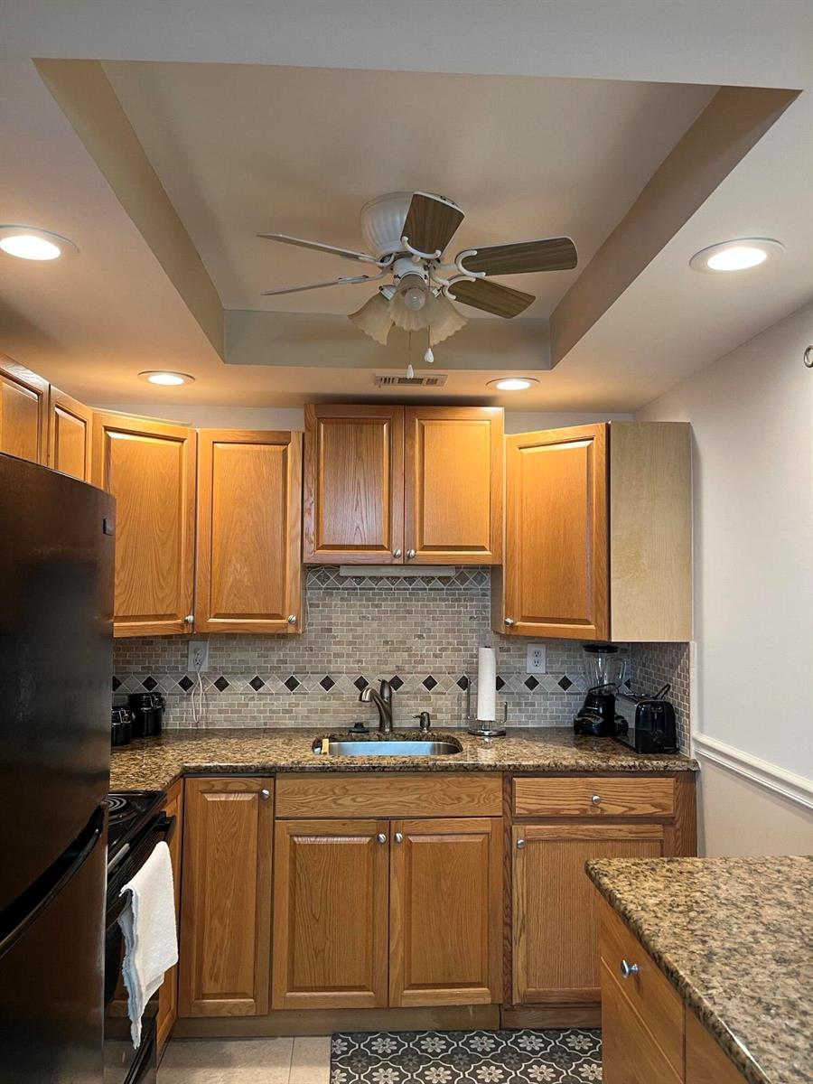 A kitchen features oak cabinets, granite countertops, a tile backsplash, and a ceiling fan — 434 Normandy # J, Delray Beach FL 33484, DELRAY BEACH, FL