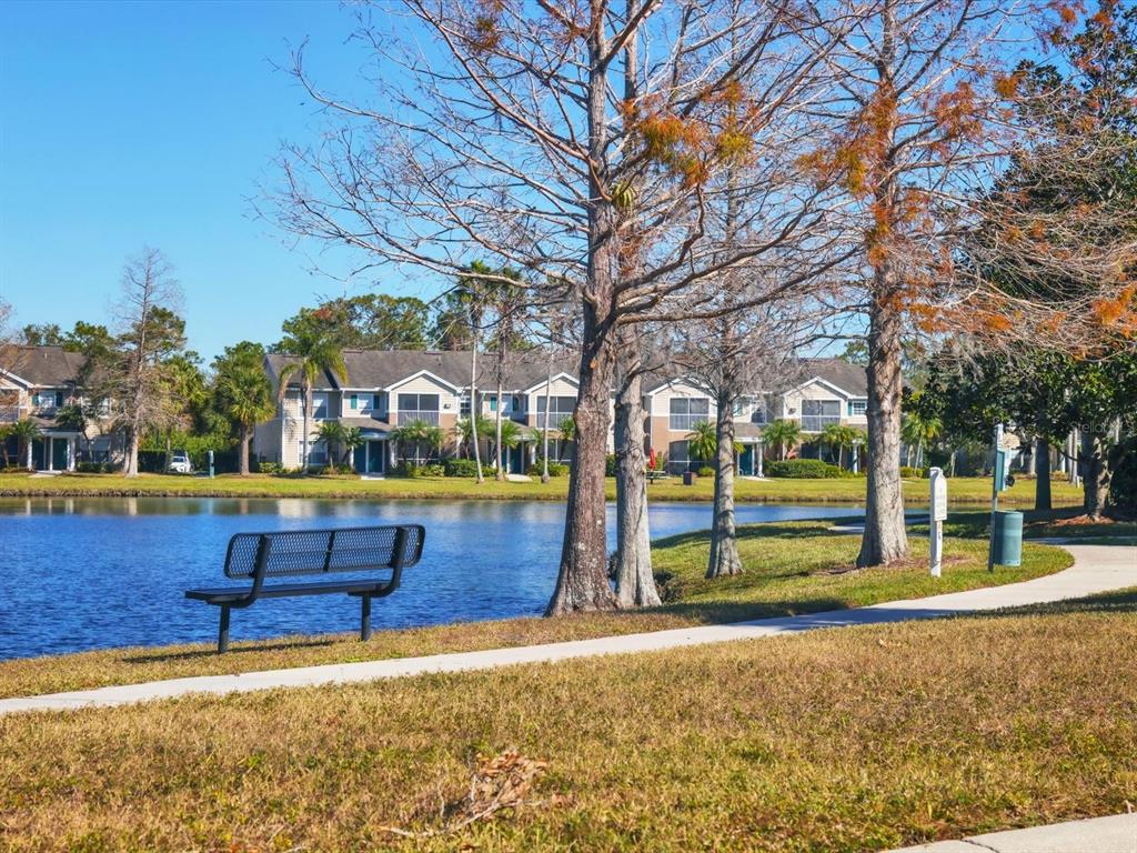 A metal bench sits on a grassy lawn beside a lake with apartment buildings behind — 8933 MANOR LOOP UNIT 105, BRADENTON, FL
