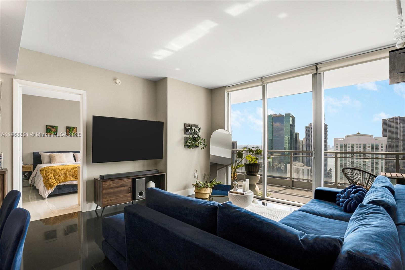 A living room with a blue sectional sofa, wall-mounted television, and a view of skyscrapers — 92 SW 3rd St # 4312, Miami FL 33130, MIAMI, FL