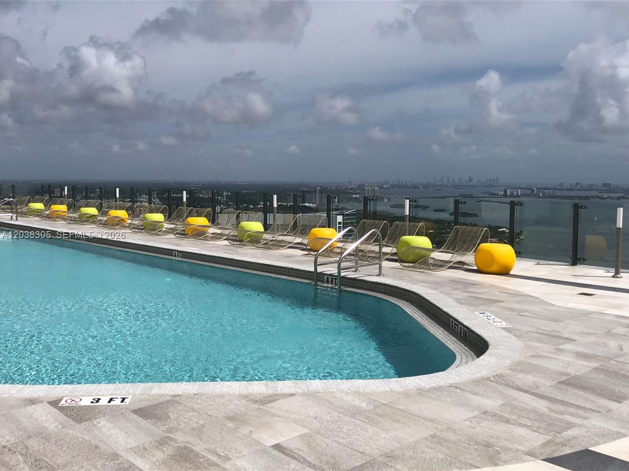 A rooftop swimming pool features lounge chairs and yellow ottomans overlooking a city skyline — MIAMI, FL