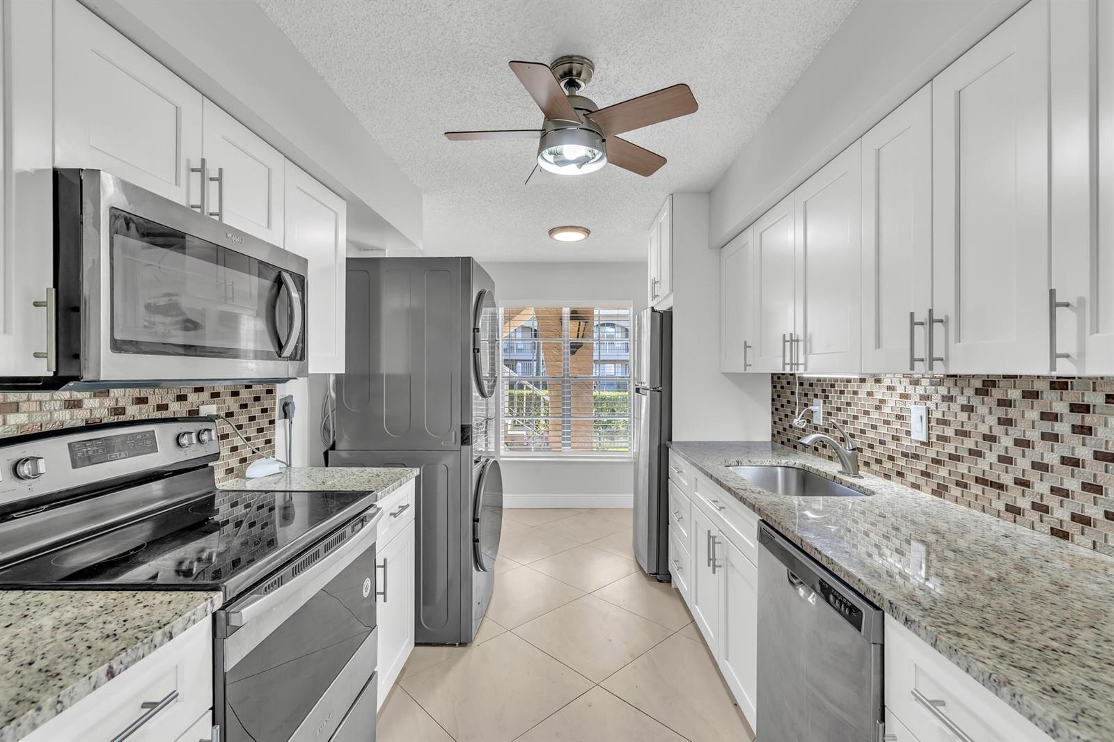 A galley kitchen features white cabinets, granite countertops, stainless steel appliances, and a stacked laundry unit — 13950 Via Flora # B, Delray Beach FL 33484, DELRAY BEACH, FL