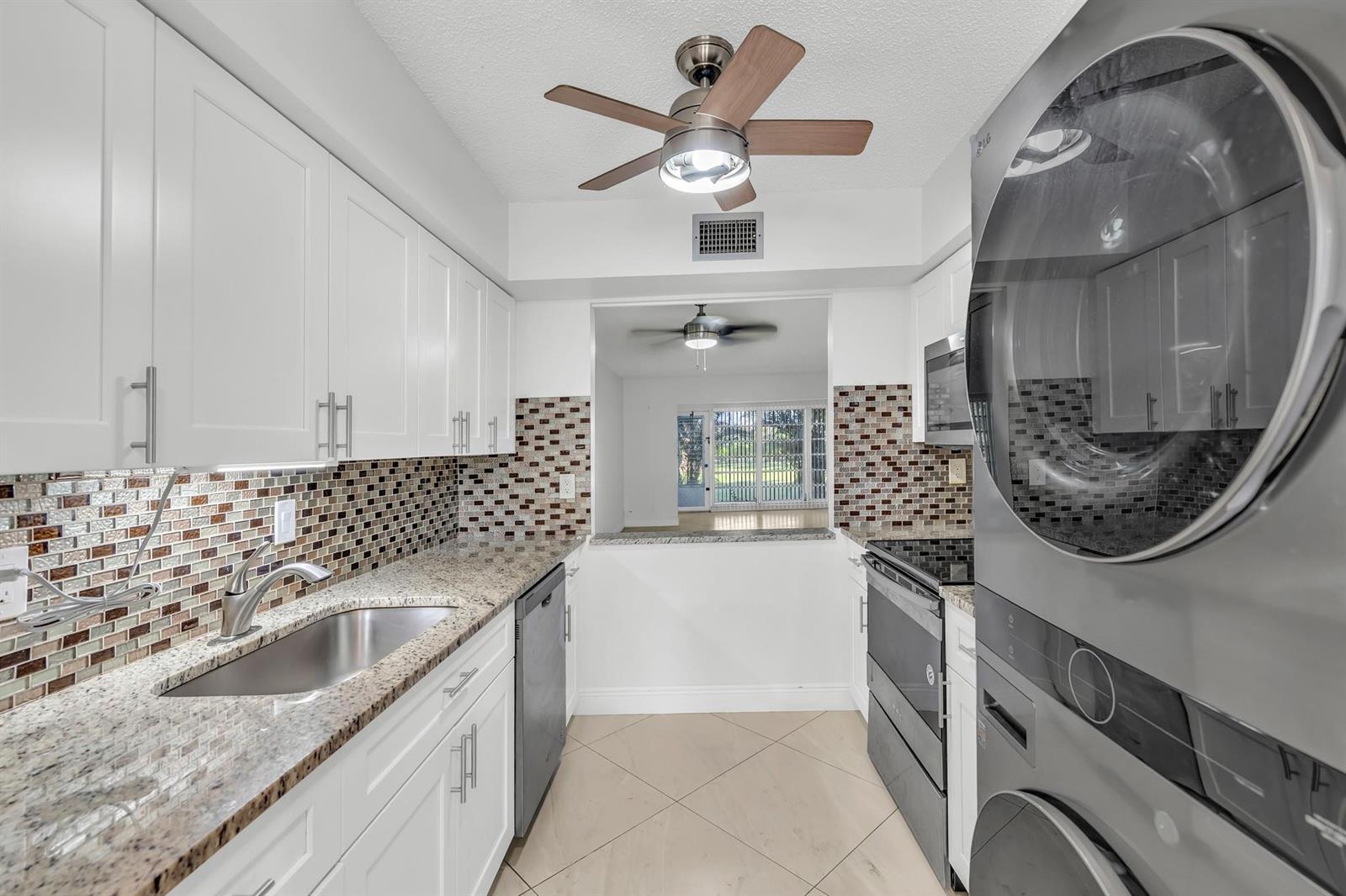 A galley kitchen features white cabinets, granite countertops, a mosaic backsplash, and a stacked washer-dryer — 13950 Via Flora # B, Delray Beach FL 33484, DELRAY BEACH, FL
